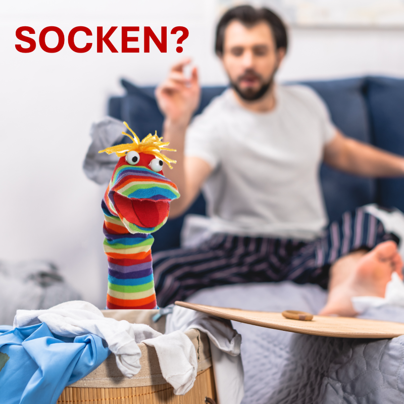 Bunter Socken-Handpuppe mit lustigem Gesicht in einem Wäschekorb, im Hintergrund ein Mann auf dem Sofa – SportBrennstoff GUTSCHEIN – Ideal für Sportler